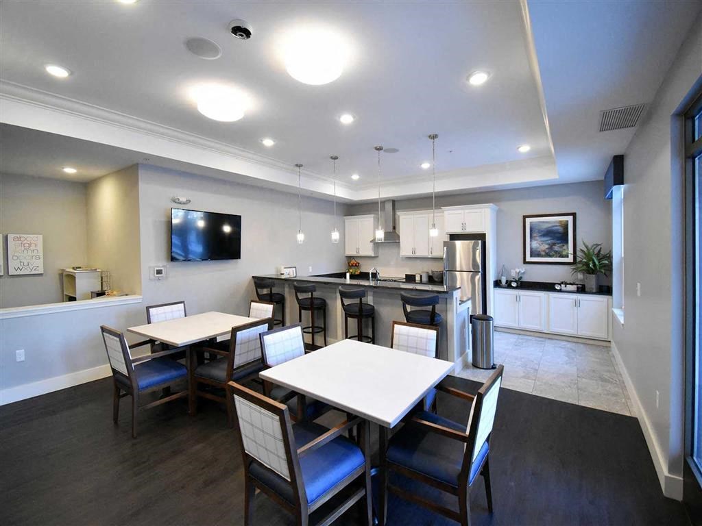 Resident lounge with modern communal kitchen with dark wood floors and gray walls at the Oaks of Shorewood Apartments. Features white cabinets, a stainless steel fridge, tables with chairs, and pendant lights.