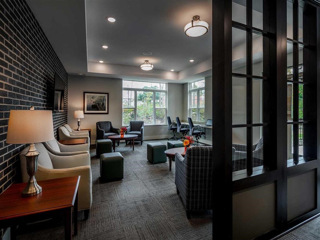 Modern lounge at the Oaks of Shorewood Apartments with soft lighting features beige armchairs, plaid chairs, and dark wood accents. Large windows provide natural light, creating a cozy atmosphere.