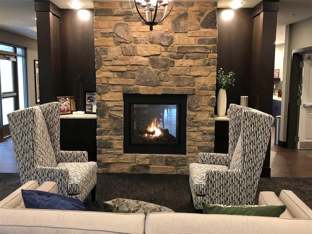 Cozy living room at the Oaks of Shorewood Apartments with stone fireplace, two patterned armchairs, a sofa with colorful cushions, and warm lighting, creating an inviting atmosphere.