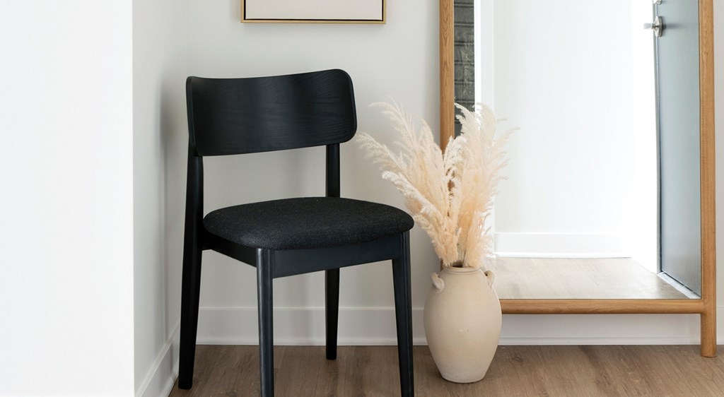 A black chair sits in front of a white vase with pampas grass.
