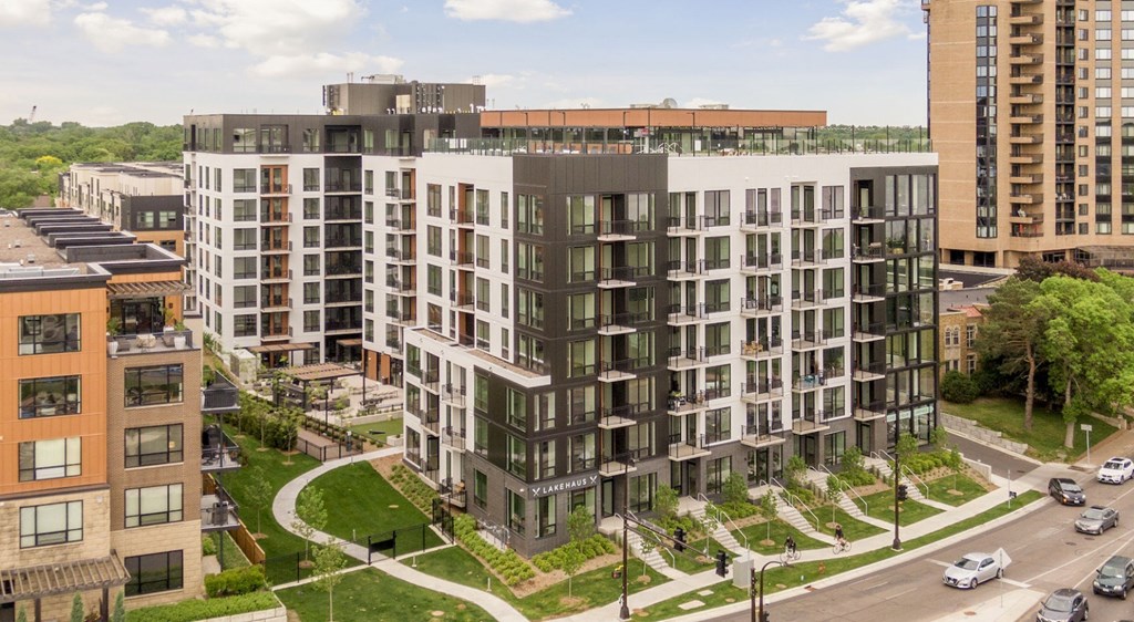 A modern multi-story building with a mix of black and white balconies is surrounded by other buildings and a street with cars.