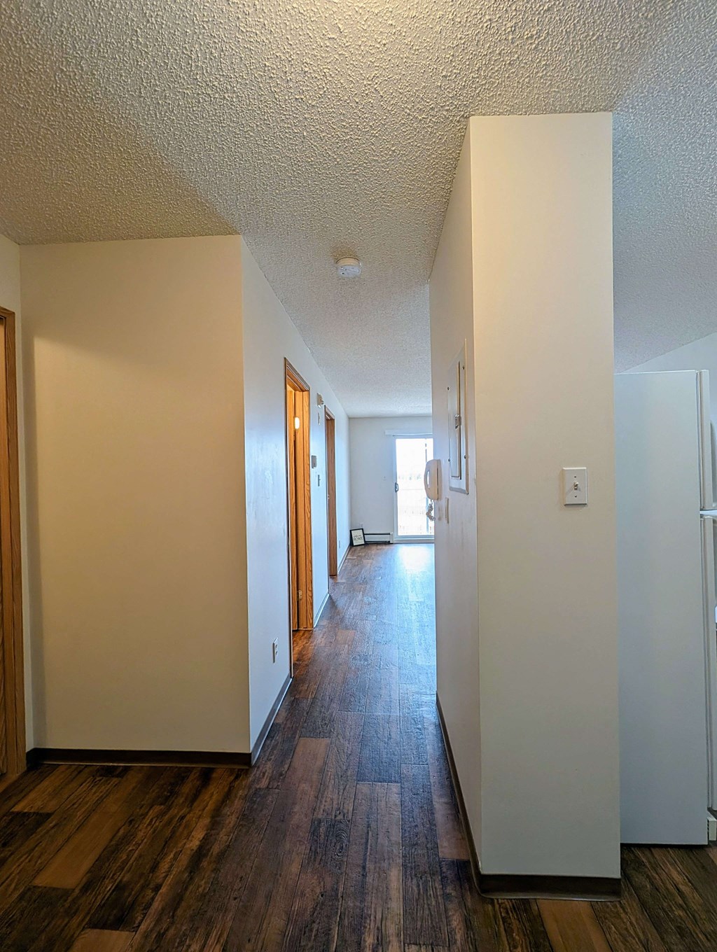 A hallway here at Aspen Terrace Apartment Homes with wooden floors and cream-colored walls, leading to a bright area with a window. Doors are on the left, creating a warm, inviting atmosphere.