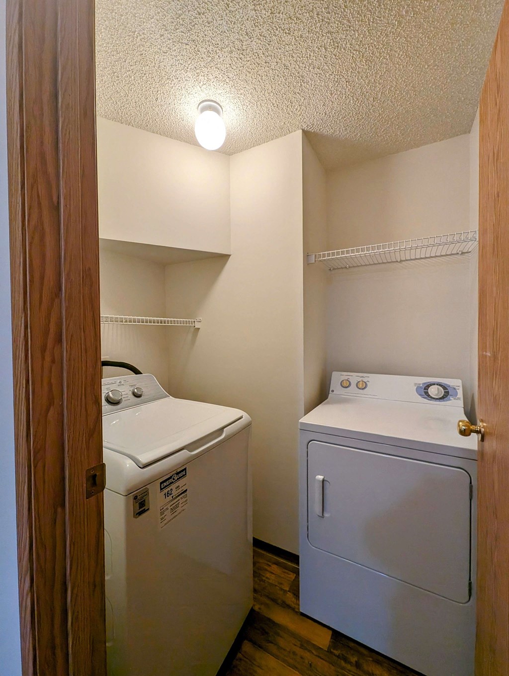 Compact laundry room here at Aspen Terrace Apartment Homes with a white washing machine and dryer under a bright ceiling light. Shelves above offer storage, complementing the wood floor.