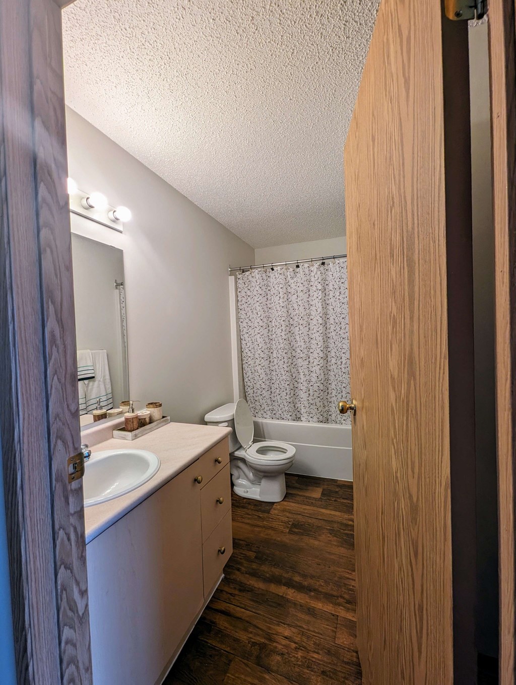 Small bathroom here at Aspen Terrace Apartment Homes with wooden floors, a white vanity with single sink, and a toilet beside a bathtub with a floral shower curtain. Warm, cozy ambiance.