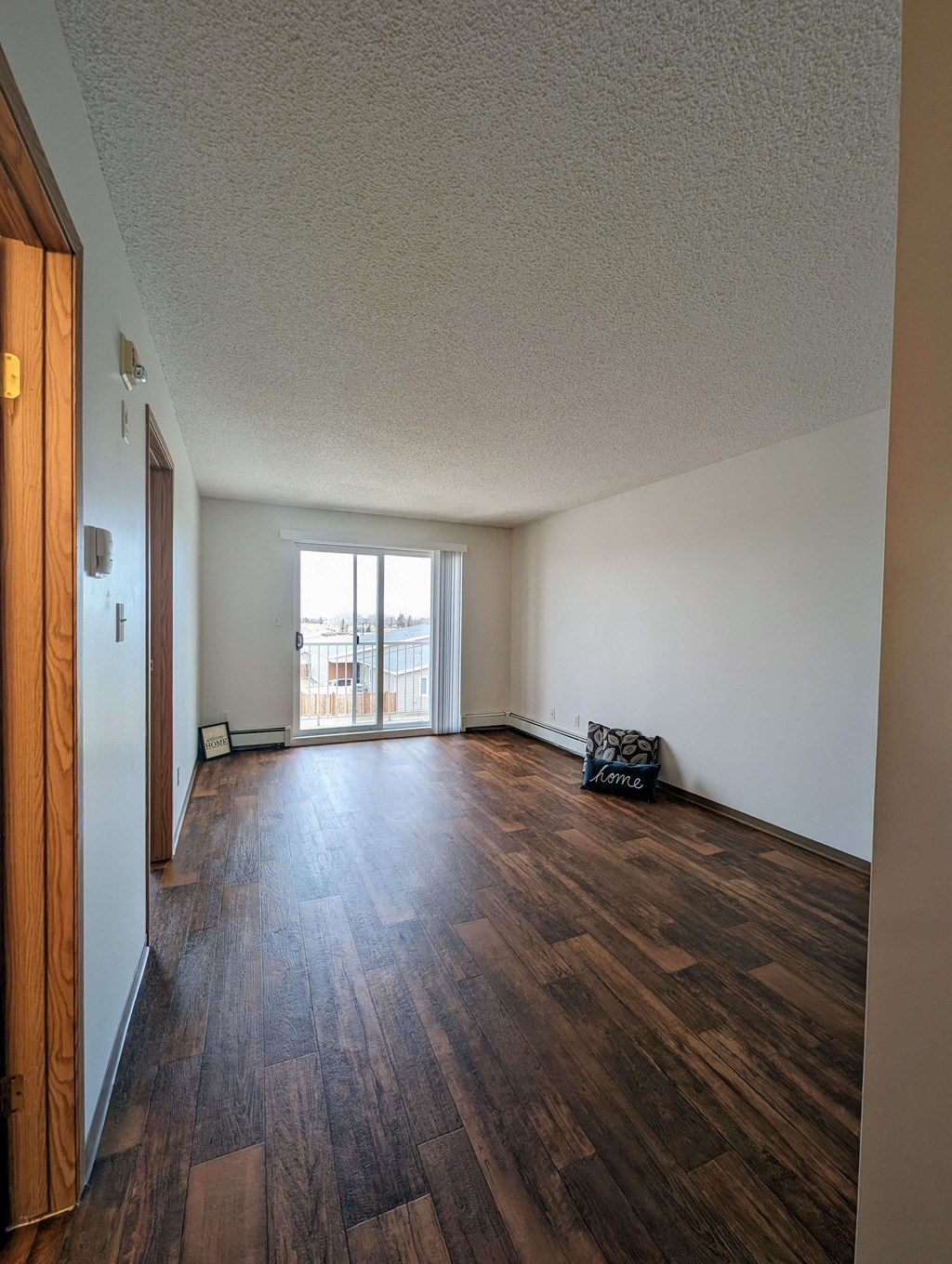 Spacious empty room here at Aspen Terrace Apartment Homes with dark wood flooring, white walls, and a sliding glass door leading to a balcony. A cushion on the floor reads "Home."