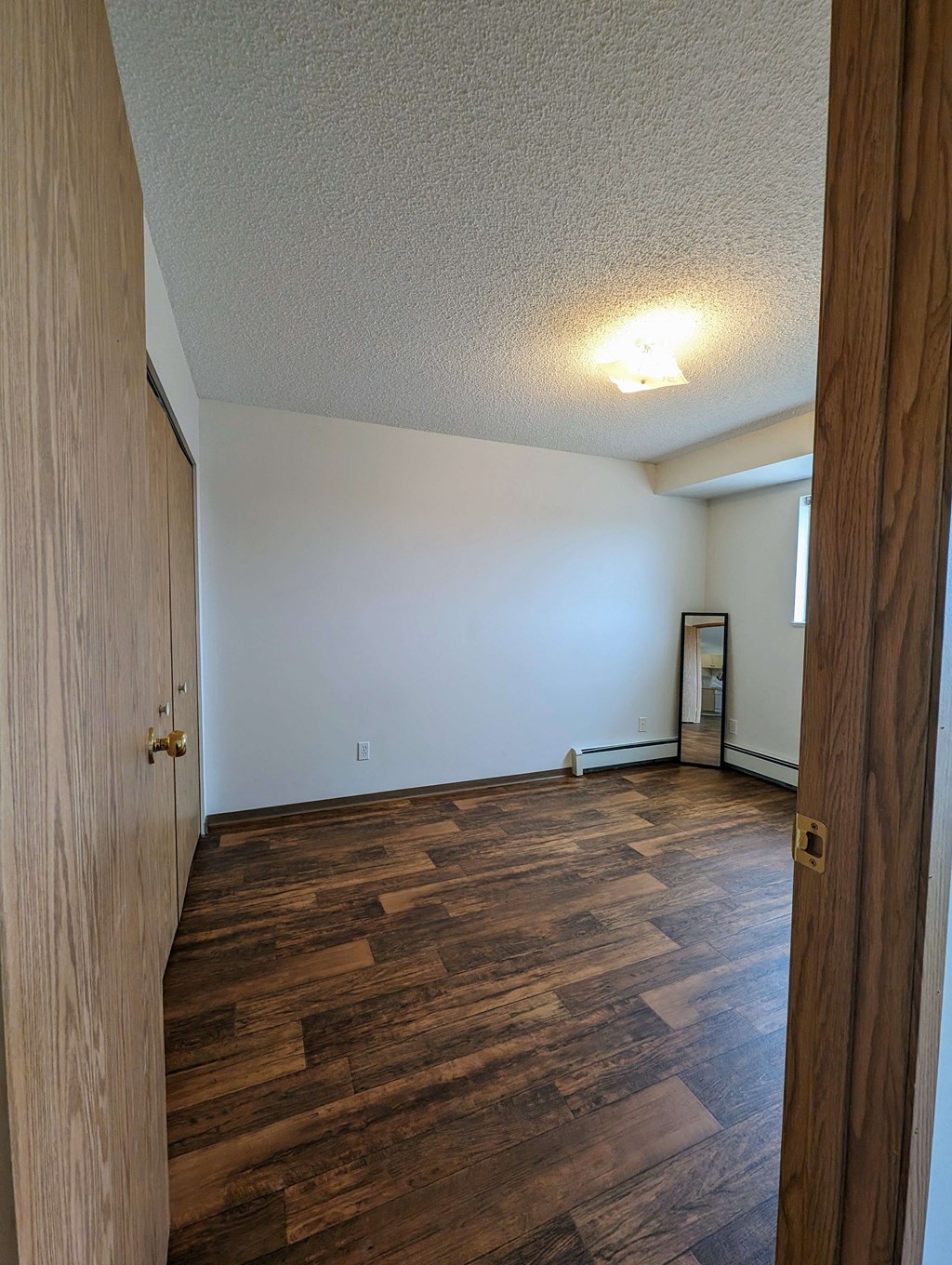 Empty bedroom here at Aspen Terrace Apartment Homes with textured white ceiling, single light fixture, wooden floor, and wood-trimmed mirror leaning against a white wall with a window. Cozy and minimalist.
