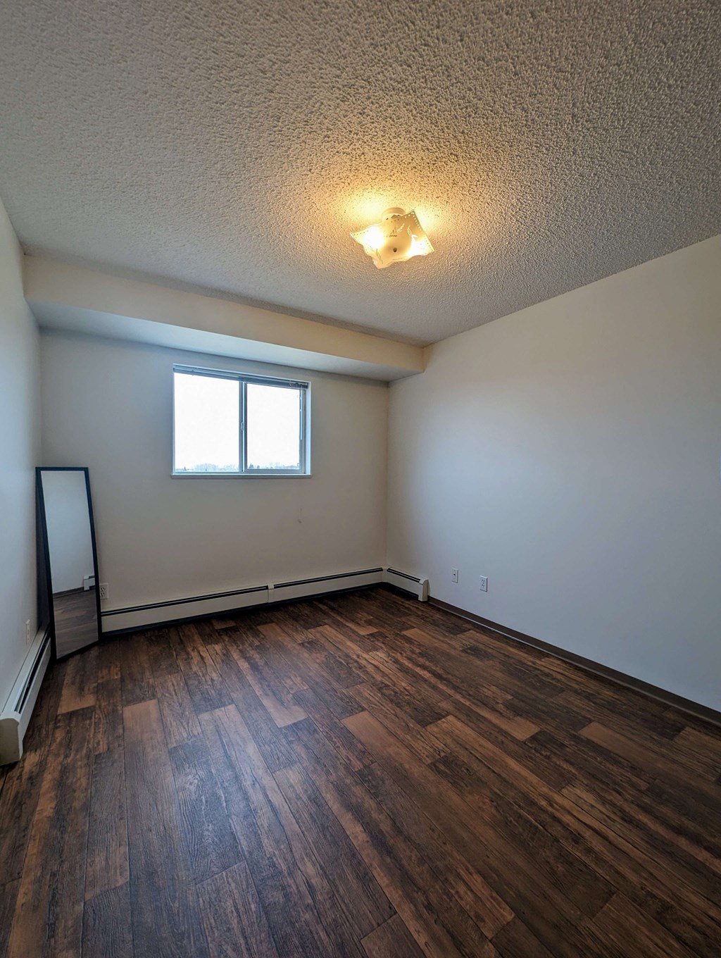 Empty bedroom here at Aspen Terrace Apartment Homes with textured white ceiling, single light fixture, wooden floor, and wood-trimmed mirror leaning against a white wall with a window. Cozy and minimalist.