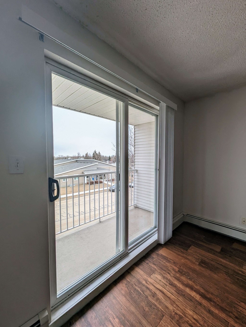 A bright, empty room here at Aspen Terrace Apartment Homes with wooden floors features a large sliding glass door leading to a balcony. The sky outside is overcast, creating a serene ambiance.