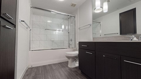 Modern bathroom here at Escape Apartments with dark wood cabinets, a large mirror, and a double sink with chrome faucets. The bathtub has a glass door and white tile walls.