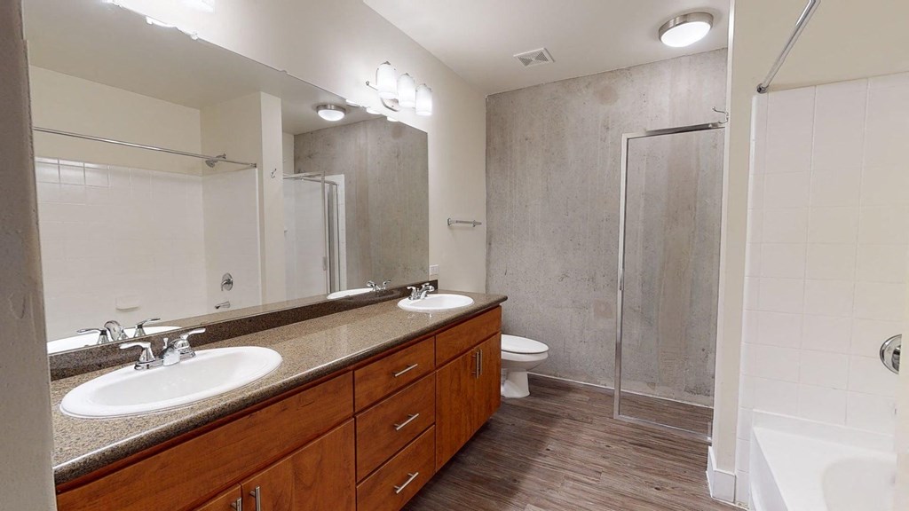 Modern bathroom here at Skyline Lofts Apartments with dual sinks on a wooden vanity, large mirror, and granite countertop. Features a glass shower, white tiles, and wood floor.
