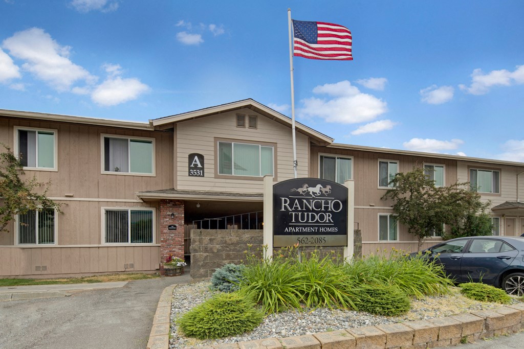 Two-story apartment building with beige siding, marked "A 3531." A U.S. flag waves above the "Rancho Tudor Apartment Homes" sign amidst landscaping.