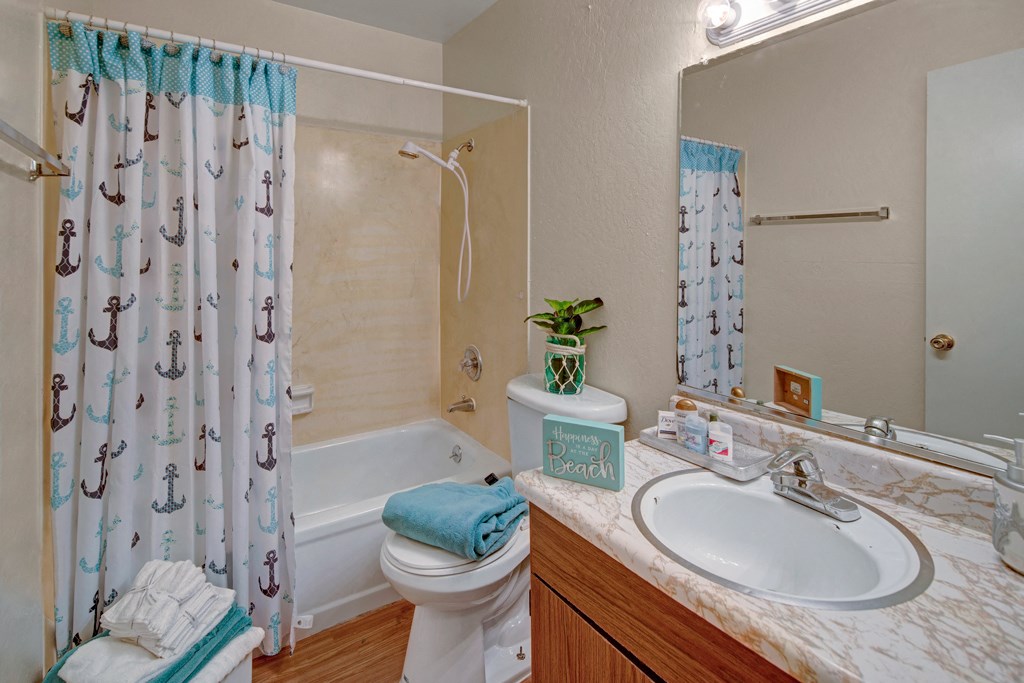 Bathroom here at Rancho Tudor Apartments with nautical-themed shower curtain featuring anchors, white bath, and sink with marble countertop. Towel, plant, and decor add a beach vibe.