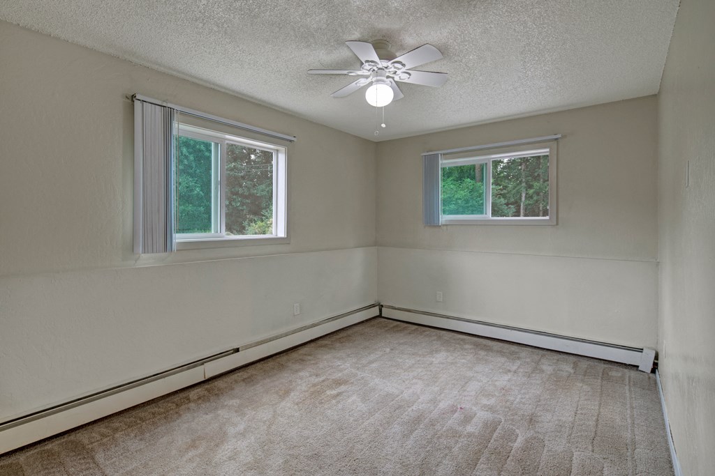 Empty bedroom here at Rancho Tudor Apartments with beige carpet and walls, featuring two large windows with vertical blinds. A ceiling fan with a light fixture hangs in the center.
