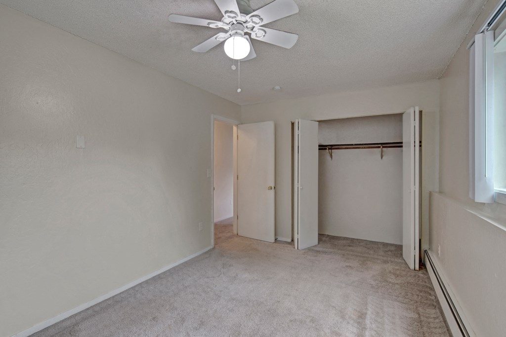 Empty bedroom here at Rancho Tudor Apartments with beige walls and carpet, featuring an open closet, a ceiling fan with light, and a window on the right letting in soft natural light.