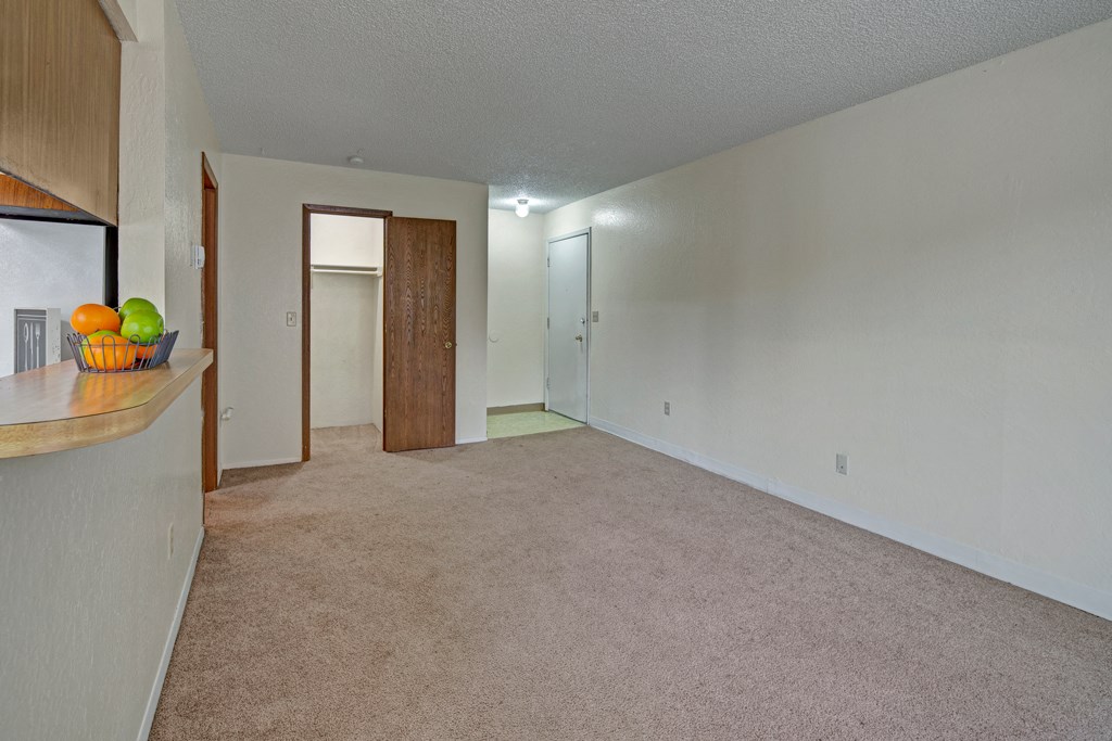 Empty living room here at Rancho Tudor Apartments with beige carpet and white walls, featuring a wooden door to a closet, a white entrance door, and a bar counter with a fruit bowl.