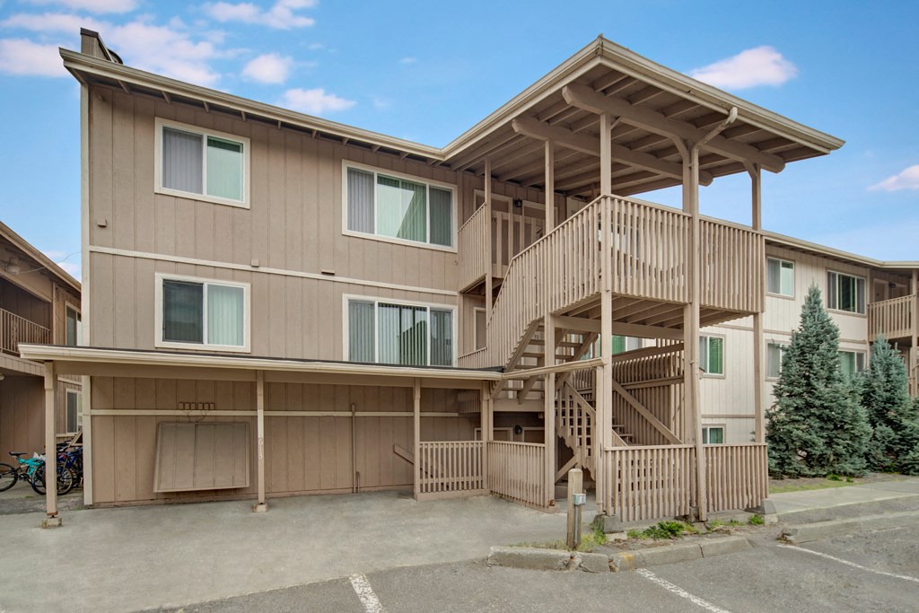 Three-story beige Rancho Tudor Apartment building with prominent wooden staircase and railings, set against a blue sky. Nearby are parked bicycles and evergreen trees.