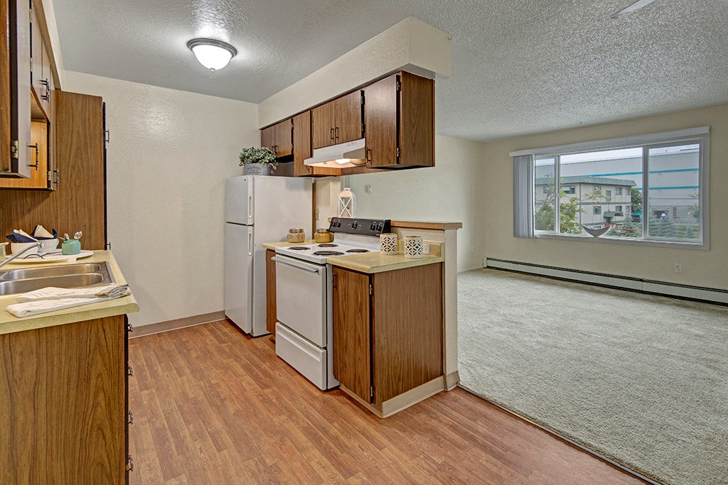Cozy kitchen here at Rancho Tudor Apartments with wooden cabinets, white appliances, and a laminate floor opens to a carpeted living area with a large window, offering natural light.
