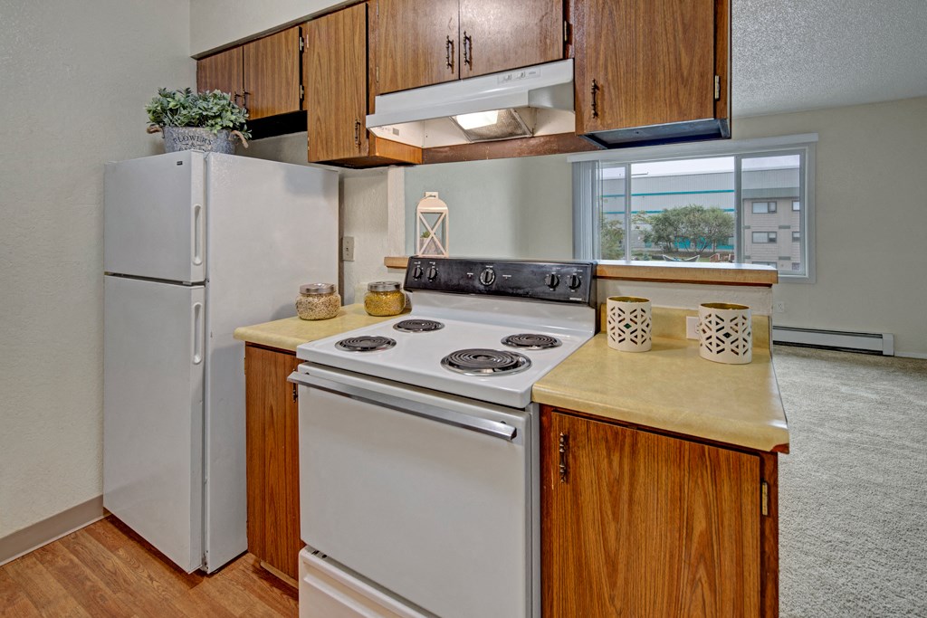 Compact kitchen here at Rancho Tudor Apartments with wood cabinets, white appliances including a stove and fridge. Decor includes plants and lanterns, with ample natural light. Cozy ambiance.