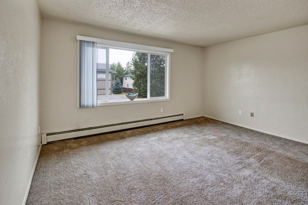 An empty beige room here at Rancho Tudor Apartments with a large window showing trees and buildings outside. The carpet is brown, the walls are plain, and there's a minimal, neutral feel.