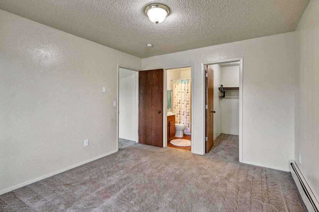 Empty carpeted room here at Rancho Tudor Apartments with beige walls, featuring two wooden doors. One door leads to a bathroom with a shower curtain; the other to a closet. A ceiling light softly illuminates the space.