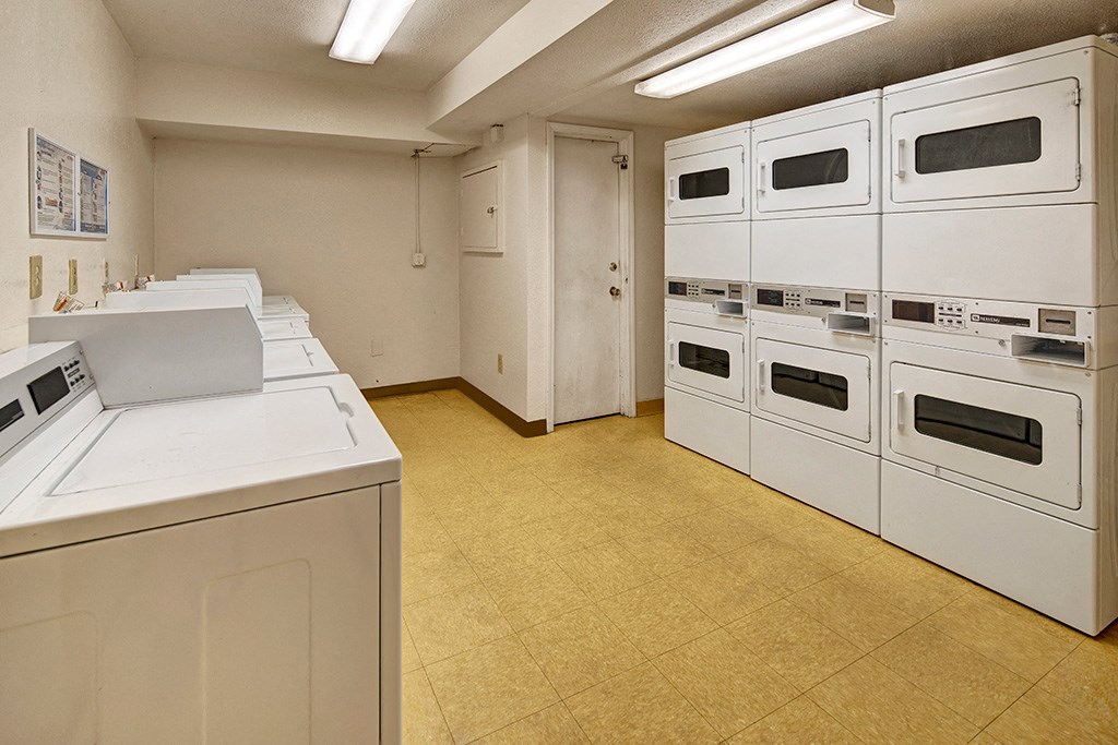 Laundry room here at Rancho Tudor Apartments with white machines and beige floor. Six stacked dryers are on the right, and five washing machines are on the left. Bright fluorescent lights illuminate the room.