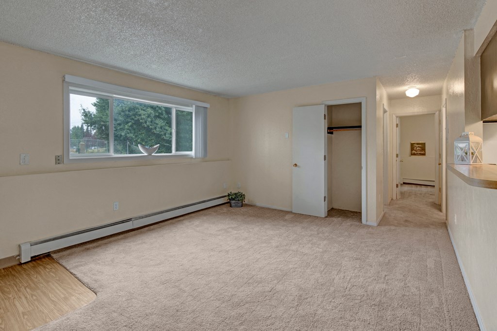 Empty beige living room here at Rancho Tudor Apartments with a large window showing greenery outside. The room features carpet, an open closet with a door, and a hallway with dim lighting. Calm and neutral ambiance.