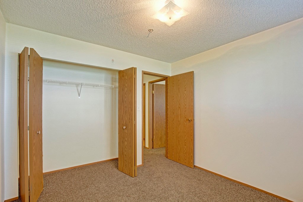 Empty bedroom here at Aspen Terrace Apartment Homes with beige walls and brown carpet. Open wooden closet and bedroom doors reveal a bright hallway. A small ceiling light softly illuminates.