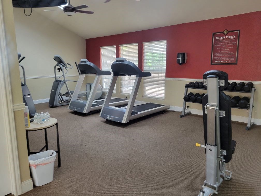 Small gym here at Trinity Place Apartment Homes with treadmills, an elliptical, and weight rack against red and beige walls. Bright light through windows, creating an inviting atmosphere.