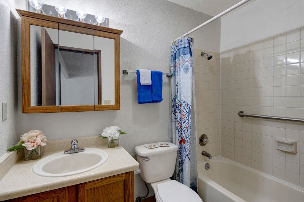 Small, tidy bathroom here at Russian Jack Apartments with a white sink, wooden cabinet, and mirror. A flower arrangement and blue towels add color. Shower has a patterned curtain.