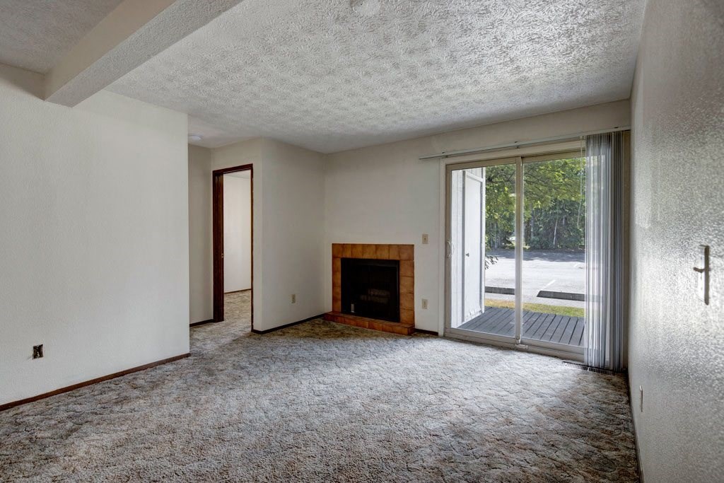 Empty living room here at Russian Jack Apartments with textured ceiling, beige carpet, and neutral walls. Features a brick fireplace, sliding glass door with vertical blinds, and natural light. Atmosphere feels calm and open.