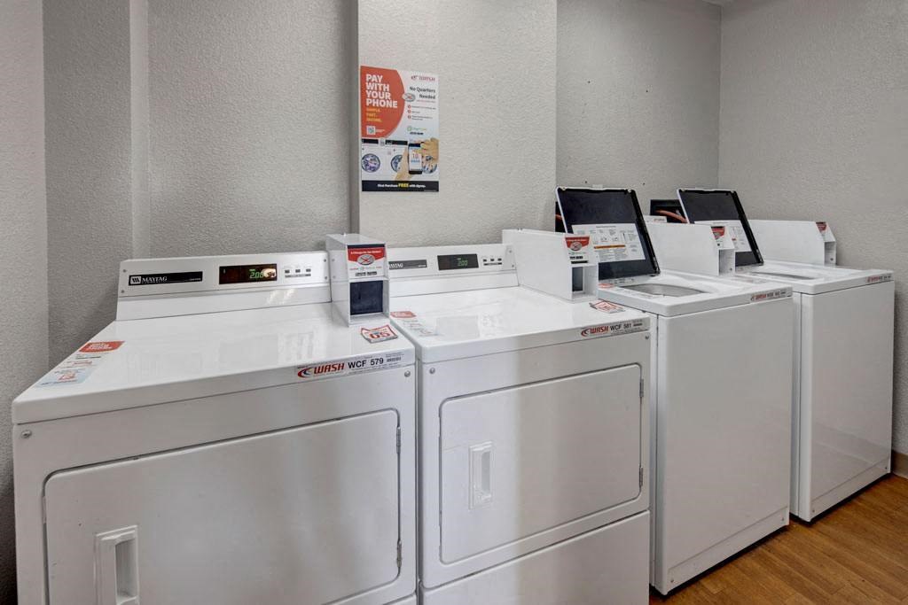 A row of white commercial washers and dryers is in a clean, gray laundry room here at Russian Jack Apartments. A poster on the wall advertises mobile payment options.