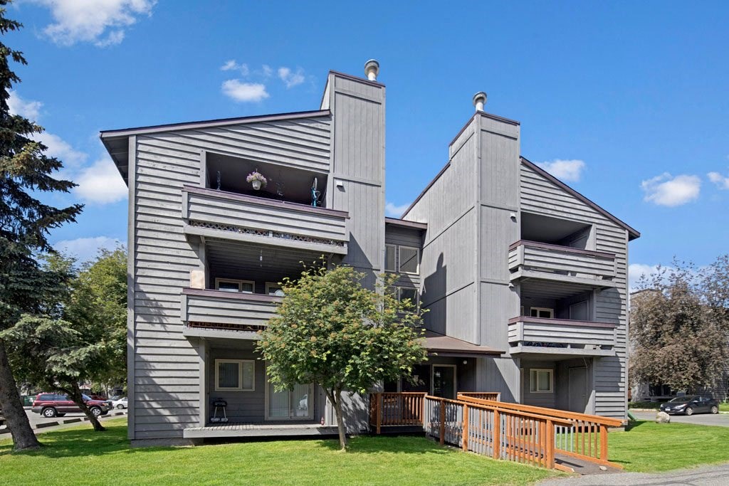 Gray three-story Russian Jack Apartment building with balconies, set against a backdrop of blue sky and trees. A wooden ramp leads to the entrance. Calm atmosphere.