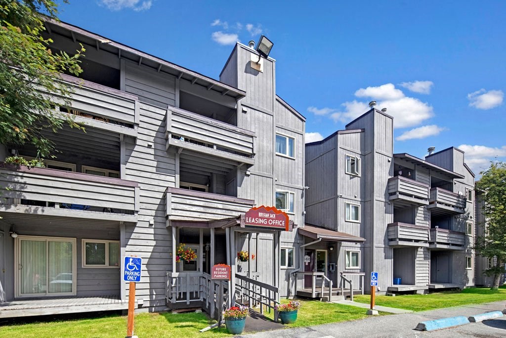 Gray, multi-story Russian Jack Apartment building with balconies under a blue sky. A leasing office sign is visible. Accessible parking signs are in front.
