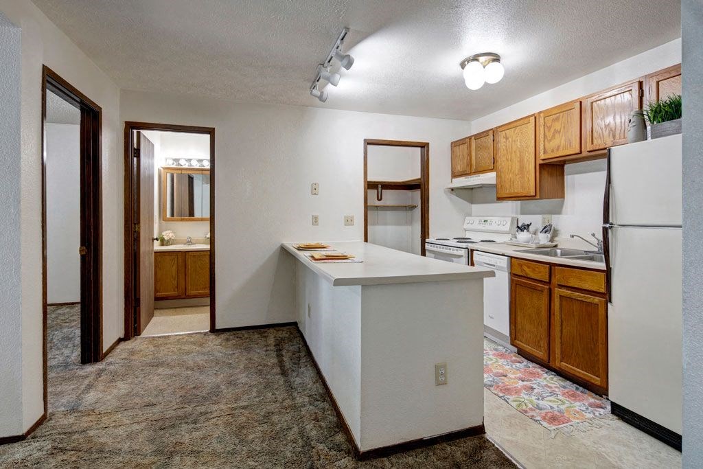 Compact kitchen here at Russian Jack Apartments with wooden cabinets, white appliances, and a floral rug. A long countertop extends into a hallway showing a lit bathroom mirror. Cozy feel.