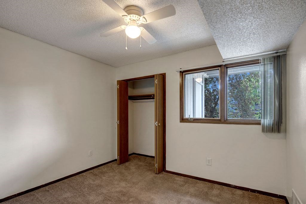 Small, empty bedroom here at Russian Jack Apartments with beige carpet, white walls, and a ceiling fan. Open closet with wooden doors and a window on the right with vertical blinds.