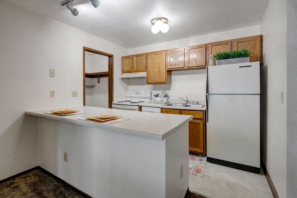 Small, cozy kitchen here at Russian Jack Apartments with overhead lighting, featuring wooden cabinets, a white fridge, stove, and sink. Simple counter with two place settings.
