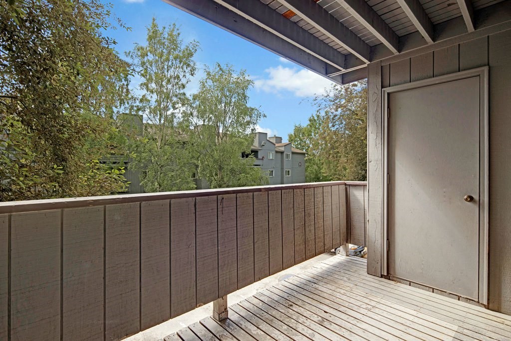 Russian Jack Apartments - Patio A wooden balcony here at Russian Jack Apartments with railing offers a view of trees and distant buildings. A closed door is on the right, under a partially covered roof, in daylight.