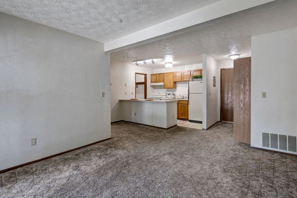 Empty living room here at Russian Jack Apartments with beige carpet, white walls, and an adjacent kitchen featuring wooden cabinets, white appliances, and a small island. Cozy and neutral.