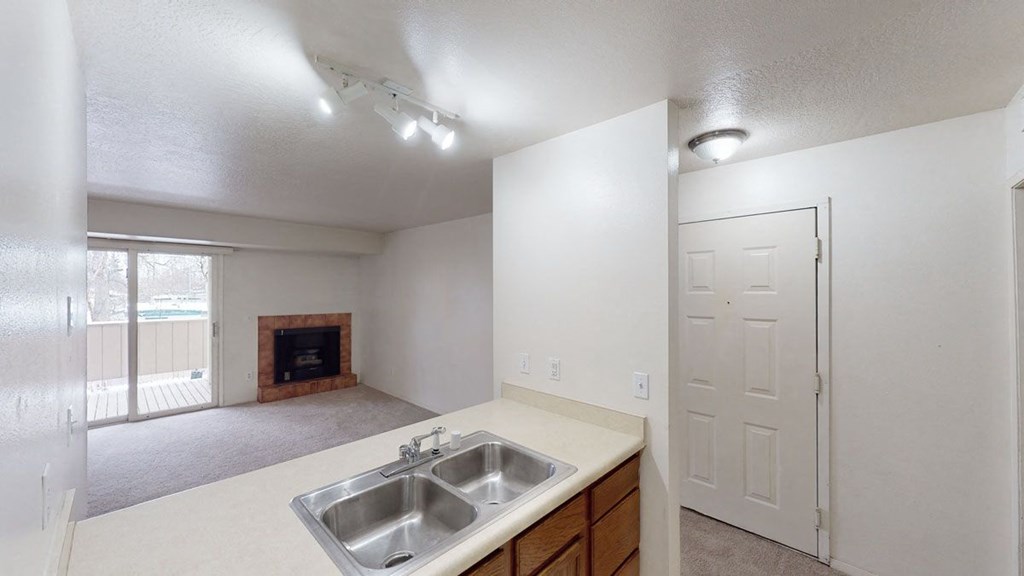 Small, empty living room here at Russian Jack Apartments with beige walls and carpet. Features a fireplace, sliding glass door to a balcony, and a kitchen view with a double sink. Neutral lighting.