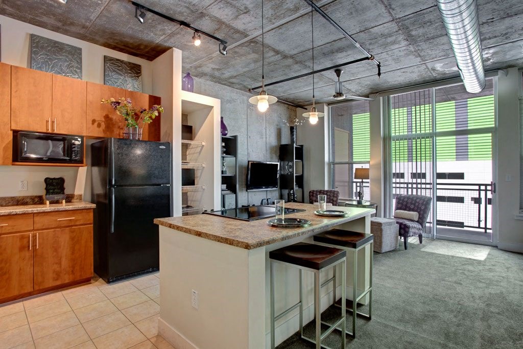 Modern industrial kitchen and living space here at Skyline Lofts Apartments with exposed ductwork and concrete ceiling. Features wood cabinets, black appliances, a central island with bar stools, and large windows offering natural light. Cozy seating area with patterned chairs and minimal decor.