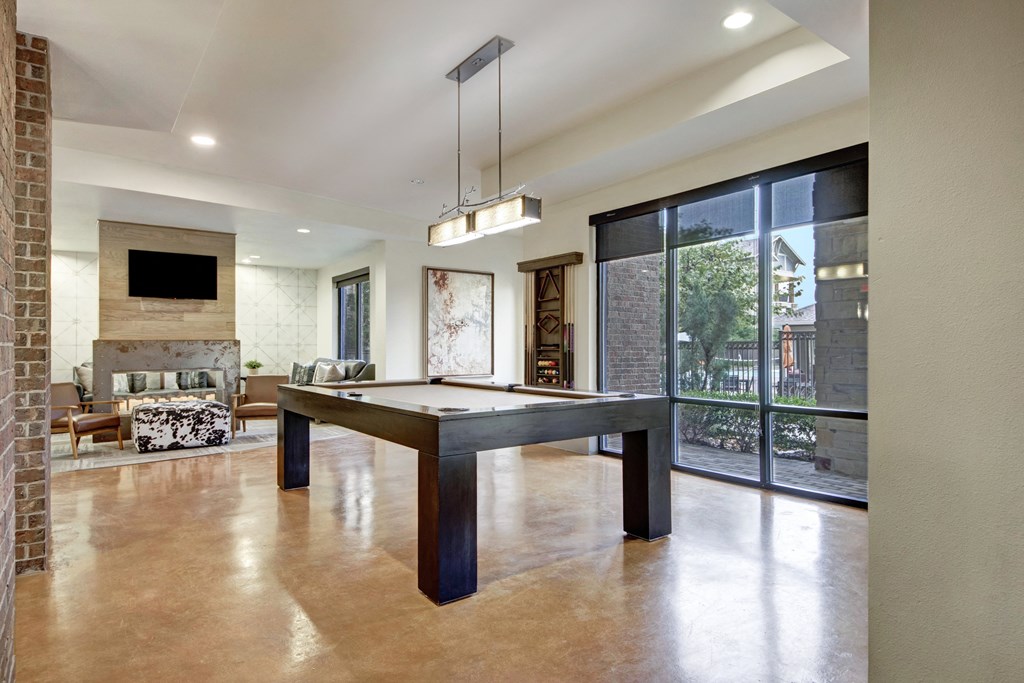 Spacious game room here at Sorrel Fairview Apartment Homes with a modern pool table centered under rectangular pendant lights. Large windows reveal a garden view. Cozy seating area with wall art and a TV in the background enhance the inviting atmosphere.