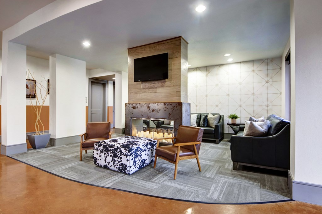 Modern lounge here at Sorrel Fairview Apartment Homes with a cozy atmosphere, featuring a central fireplace, two leather chairs, a cowhide ottoman, and a TV above. Neutral tones and soft lighting create a calming environment.