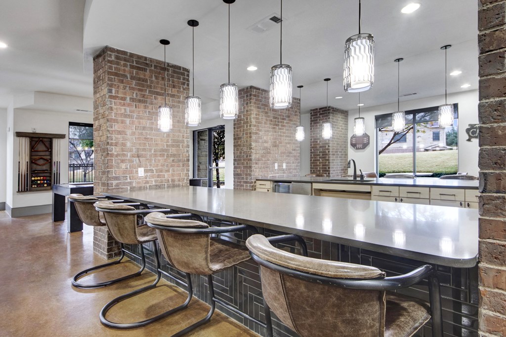 Modern kitchen here at Sorrel Fairview Apartment Homes with brick accents. Long gray countertop with five brown chairs. Eight pendant lights hang above, creating a warm, inviting ambiance.