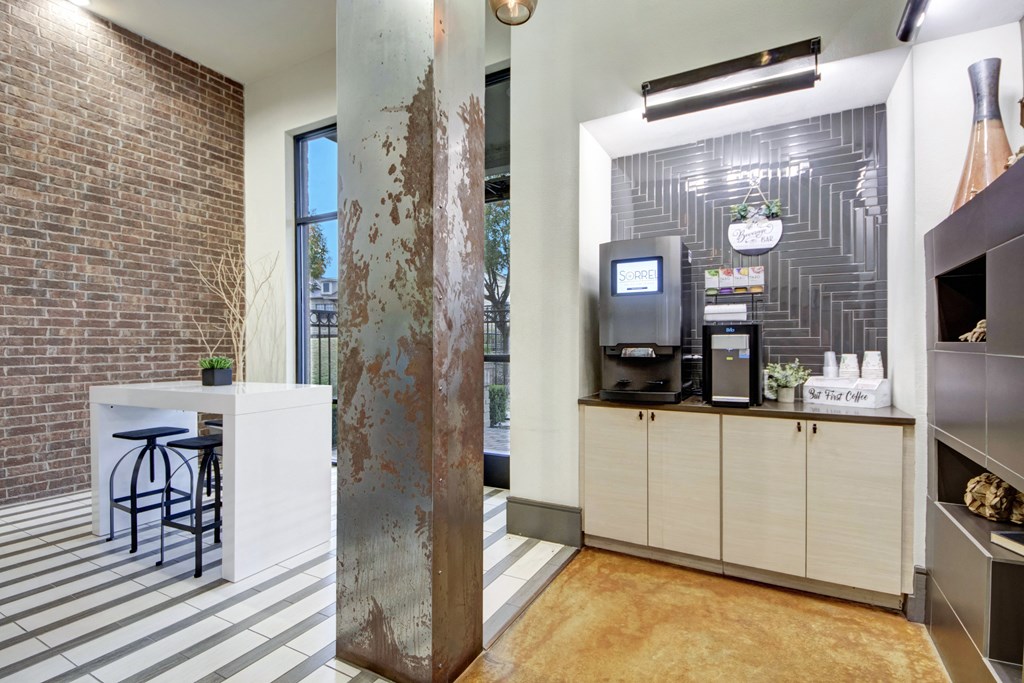 Modern coffee station here at Sorrel Fairview Apartment Homes in a cozy room with brick and tile walls, featuring a sleek coffee machine, cups, and a nearby white table with high stools.