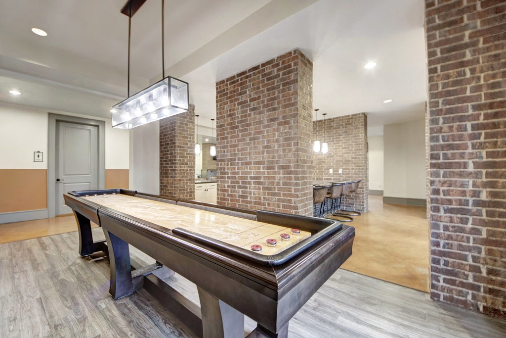 Spacious game room here at Sorrel Fairview Apartment Homes with a shuffleboard table, brick walls, and modern lighting. In the background, a counter with bar stools adds a social feel.
