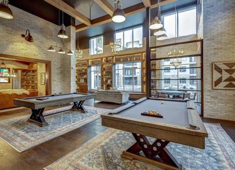 A game room here at Alden at Cedar Park with two pool tables, patterned rugs, tall windows, brick walls, wood beams, and warm pendant and chandelier lighting in a spacious lounge.