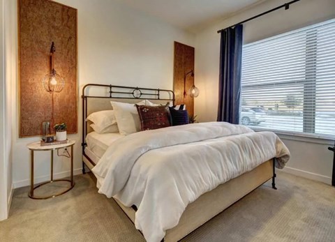 A bedroom here at Alden at Cedar Park with a black metal bed frame, white bedding, dark accent pillows, blue curtains, round gold side table, and warm wall panels with hanging lights.