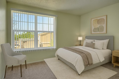 A calm, inviting bedroom here at Avalon Springs with soft neutral walls and plush carpet, featuring a comfortable bed with light bedding, a bedside lamp, and natural light filtering through a large window with vertical blinds for a bright and restful atmosphere.