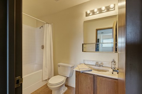A clean, modern bathroom here at Avanti Townhomes with a bathtub-shower combo, neutral walls, and a wood-tone vanity with a light countertop, featuring a wall-mounted mirror, bright lighting, and a functional, polished layout.