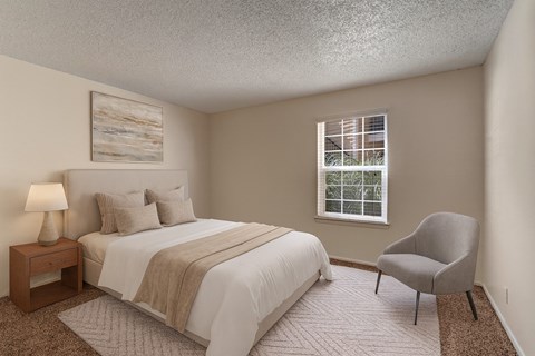 A bright bedroom here at The Bradford with beige carpet, soft neutral walls, and a large window with blinds allowing natural light to fill the space. A neatly staged bed with neutral bedding and a simple accent chair create a calm, comfortable, and inviting atmosphere.